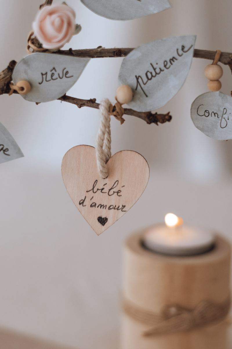 Cœur en bois gravé « bébé d'amour » suspendu à l'arbre, entre les feuilles « rêve » et « patience », devant une bougie allumée — Entre Deux