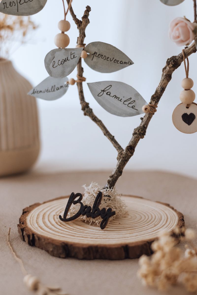 Le mot « Baby » découpé dans le bois au pied de l'arbre des valeurs, posé sur son rondin — Entre Deux