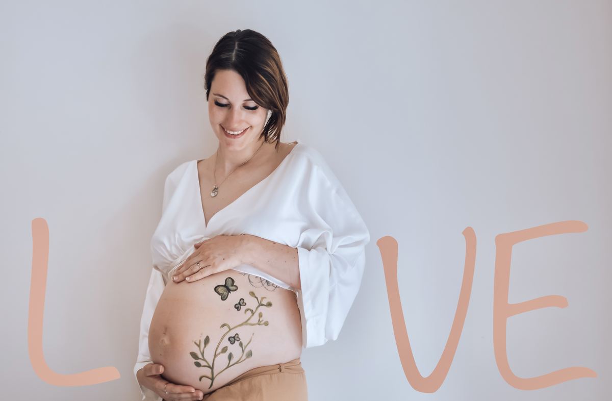 La future maman entre les lettres L et VE peintes en rose, son ventre peint formant le O de « love » — Entre Deux