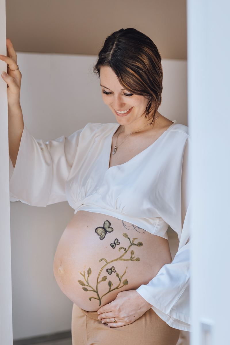 Ventre peint d'un papillon et de branches fleuries, la future maman appuyée contre le mur — Entre Deux