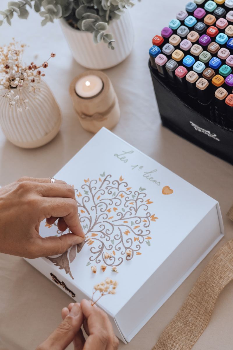 Mains en train de colorier l'arbre dessiné sur le couvercle du coffret, les feutres posés à côté — Entre Deux