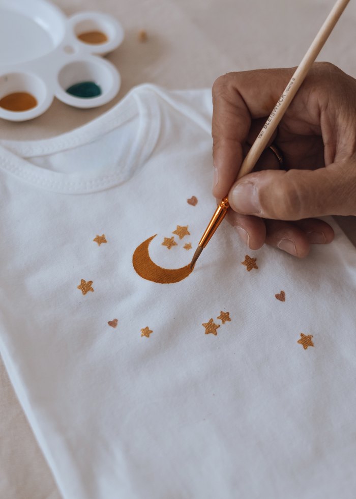 Pinceau doré traçant une lune et des étoiles sur un body de bébé pendant un atelier — Entre Deux, Enghien